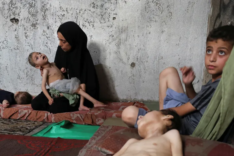 Samah Matar, una madre palestina desplazada, sostiene a su hijo desnutrido, Ameer, que sufre de parálisis cerebral, mientras su otro hijo, Youssef, también desnutrido y con parálisis cerebral, yace en un colchón en una escuela de la ciudad de Gaza. [Mahmoud Issa/Reuters] AL JAZEERA